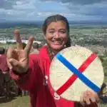 Abby Lampe holds up two fingers and a large wheel of cheese standing on top of a hill with a crowd of people behind her.