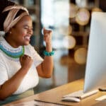 Business, night and black woman with achievement, fist and computer with email, celebration and success. African American female leader, manager and ceo in workplace, profit growth and website launch