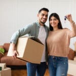 House Ownership. Young Couple Showing Keys And Holding Cardboard Box, Cheerful Guy And Lady Hugging After Moving In New Apartment Standing In Living Room. Insurance, Real Estate, Mortgage Concept