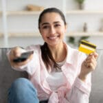 Portrait of cheerful young woman relaxing on comfortable couch at home, holding tv remote control and a golden credit card, changing channels, spending weekend and resting, ready to order