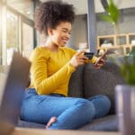 Young happy mixed race woman using a credit card and phone alone at home. Cheerful hispanic female with a curly afro making an online purchase with a debit card and cellphone while sitting on the couch at home