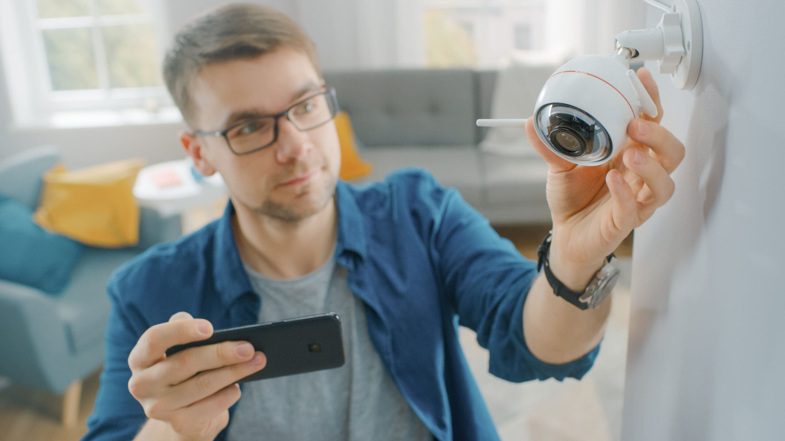 Young Man in Glasses Wearing a Blue Shirt is Adjusting a Modern Wi-Fi Surveillance Camera with Two Antennas on a White Wall at Home. He's Checking the Video Feed on his Smartphone.