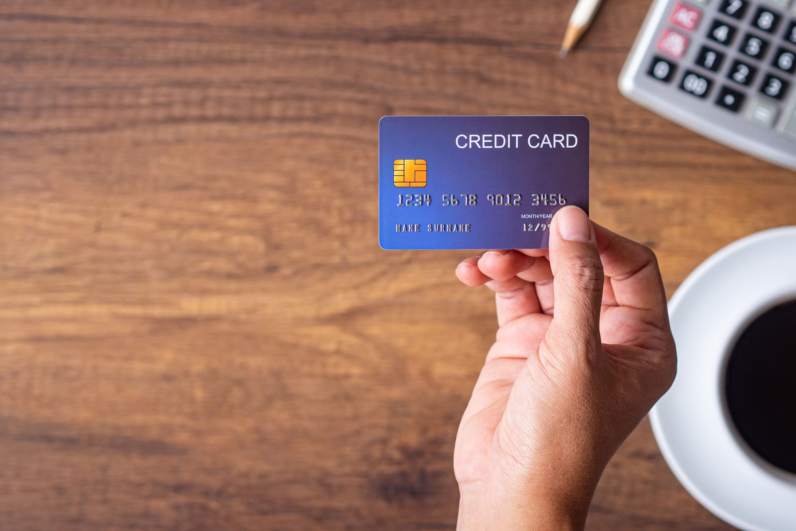 Close-up of hand holding a blue credit card with a white coffee cup, pencil, and calculator on wooden desk background. Selective focus. Space for text. Business and payment concept.