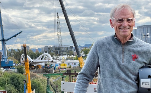 Earl Blumenauer, smiling in a gray sweater with a red bicycle pin, with the span of a new Portland bridge in the background.