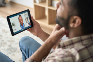 photo - Man at Home Holding Device on Telehealth Appointment with Doctor