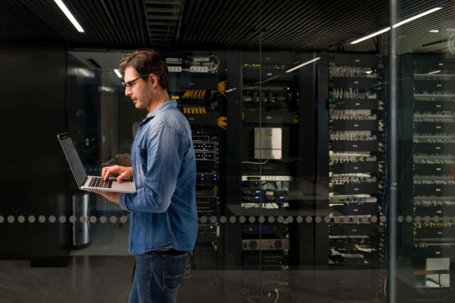 photo - Technician Working on Laptop and Walking by Server Network