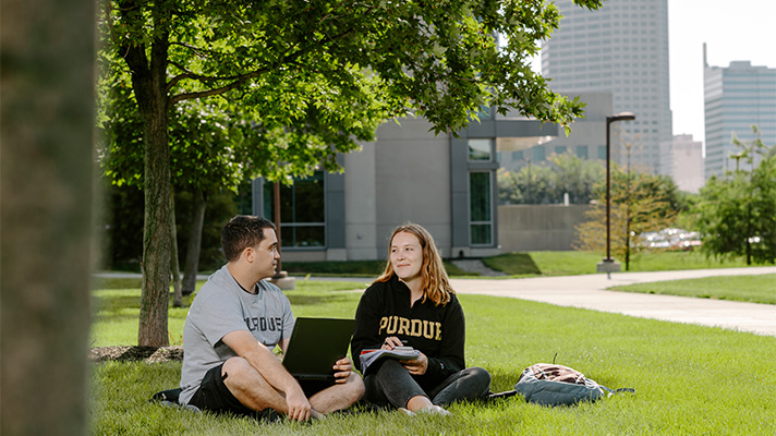 One man and one woman wearing Purdue shirts sitting beneath a shade tree on a college campus with a city in the background.