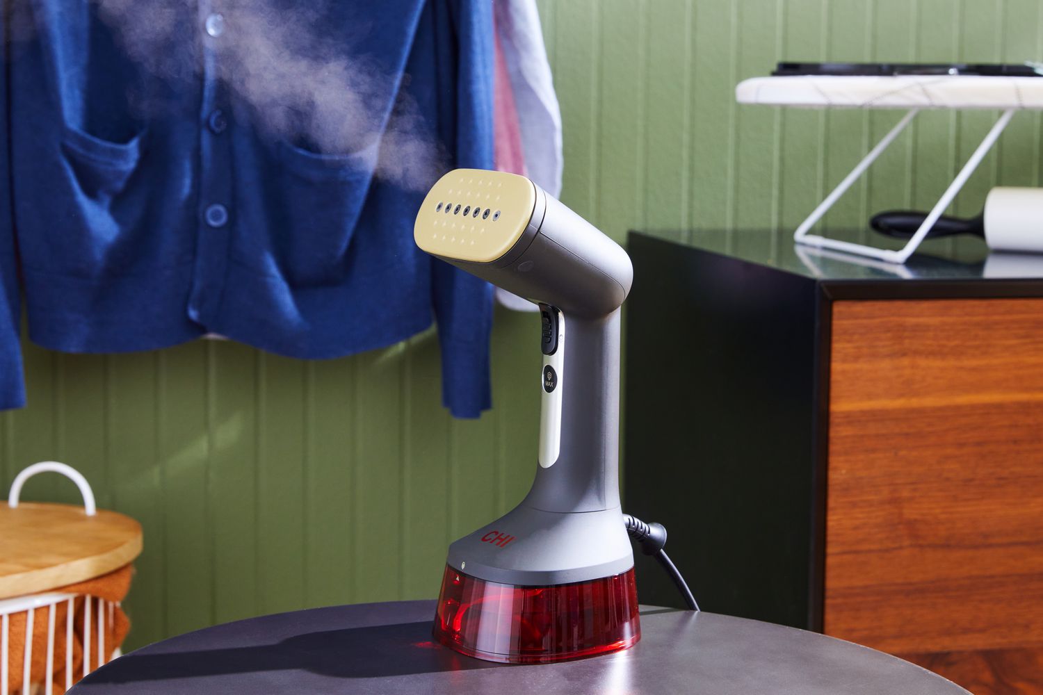 The CHI Handheld Garment Steamer on a table with steam coming out and clothes hanging in the background. 