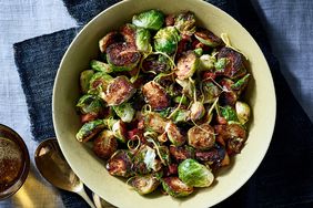 Top View of Roasted Brussels Sprouts With Pancetta Served in a White Bowl on Top of Blue Fabric, with Metal Spoon and Small Metal Bowl of Peppered Oil to the Left