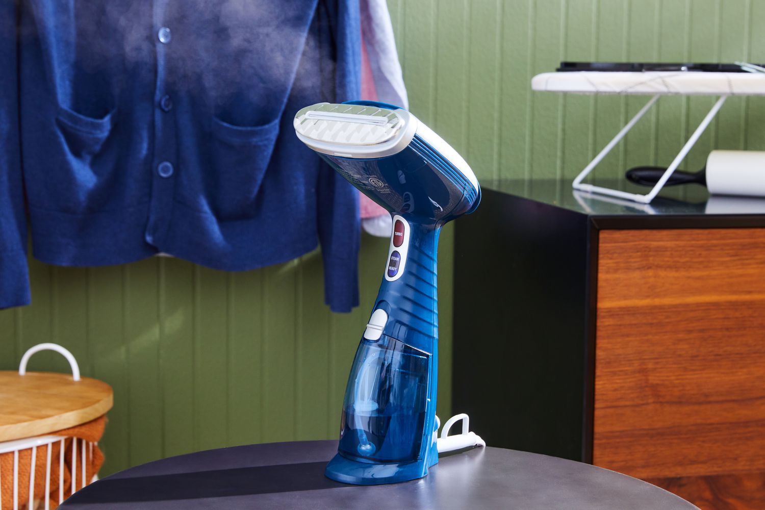 The Conair GS38R Handheld Garment Steamer on a table with steam coming out and clothes hanging in the background.