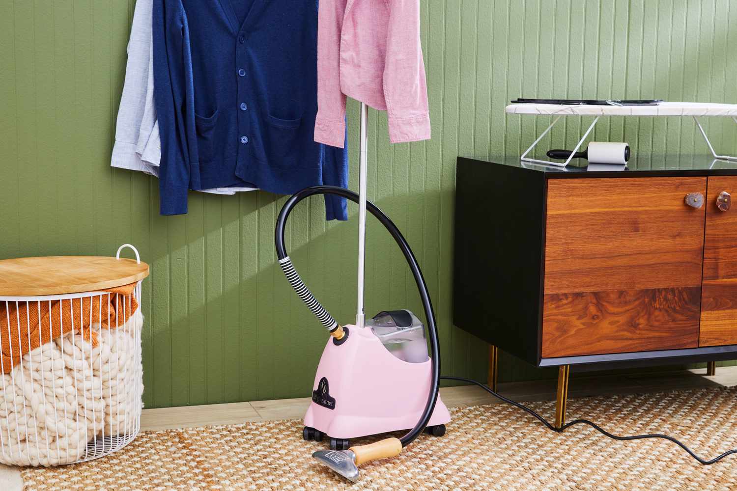 The Jiffy J-2000 Floor Standing Fabric Steamer sitting in a green room with clothes hanging on the wall.