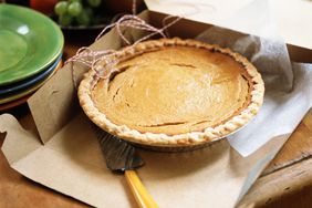 Pumpkin Pie With a Serving Knife in an Open Box Next to a Stack of Colorful Plates and a Fruit Basket