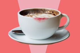 White coffee mug full of coffee with red lipstick stain on the rim, sitting on a white saucer with a metal spoon