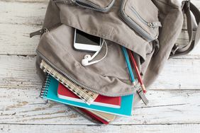 Overhead photo of gray backpack on a wooden surface, filled with notebooks, pencils, a white phone, and white headphones