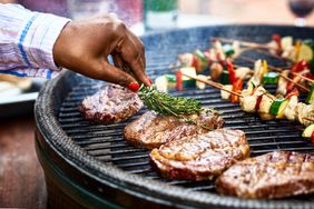 Steaks being seasoned with herbs as they cook on an outdoor grill next to shish kabobs