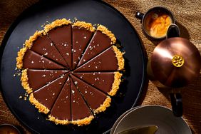 A sliced chocolate ganache tart served on a black dish sits next to a cup of coffee and french press.