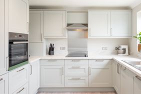A general interior view of a newly fitted sage green, grey shaker style kitchen with stainless steel handles, extractor fan hood, splash back, oven, knife block, food mixer, potted plant, recessed ceiling spot lights and marble effect worktop within a home