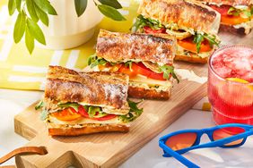 Fancy tomato sandwiches on a wooden board displayed on a table with fruit juice and sunglasses.