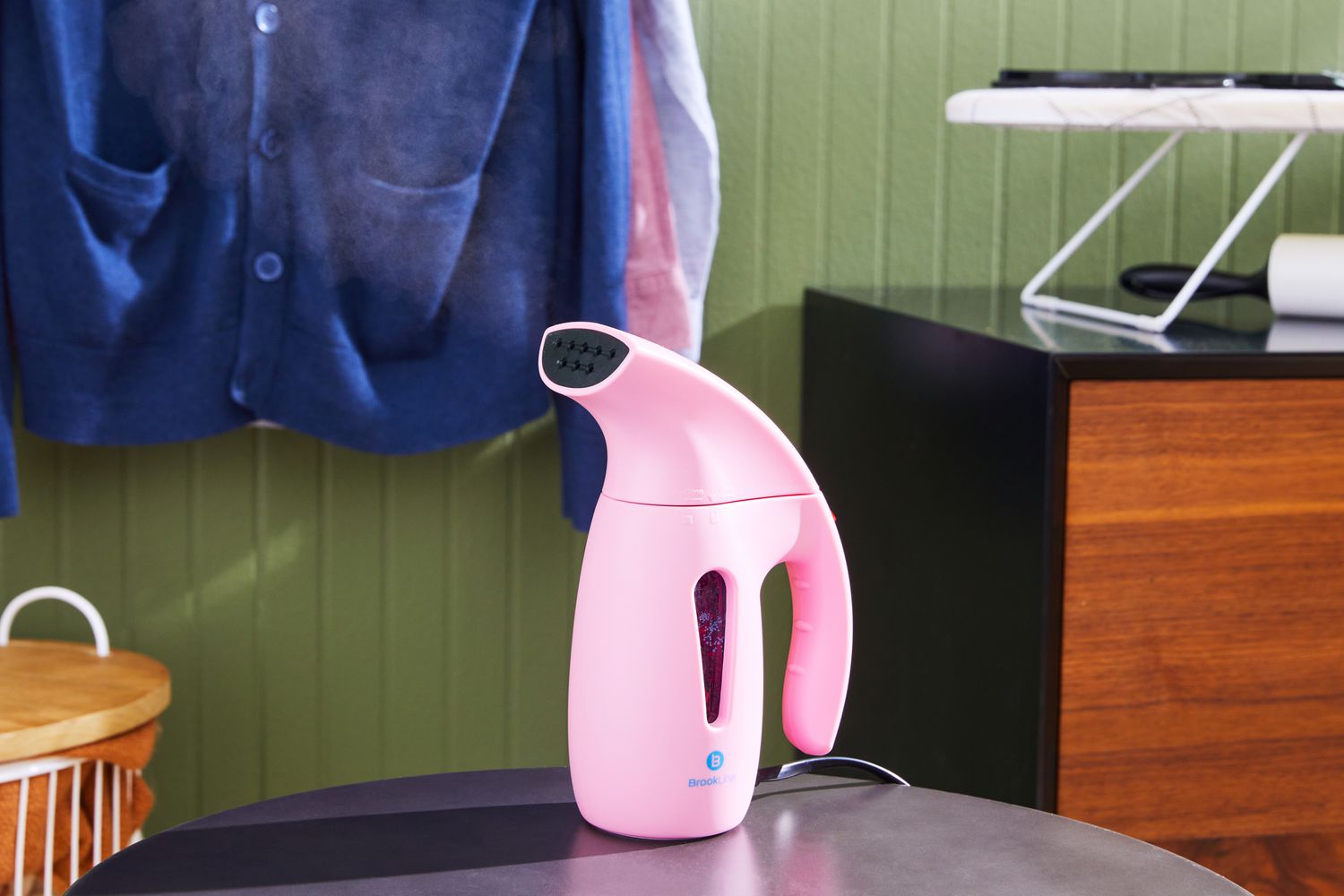The Brookline Handheld Garment Steamer on a counter with steam coming out and clothes hanging in the background. 
