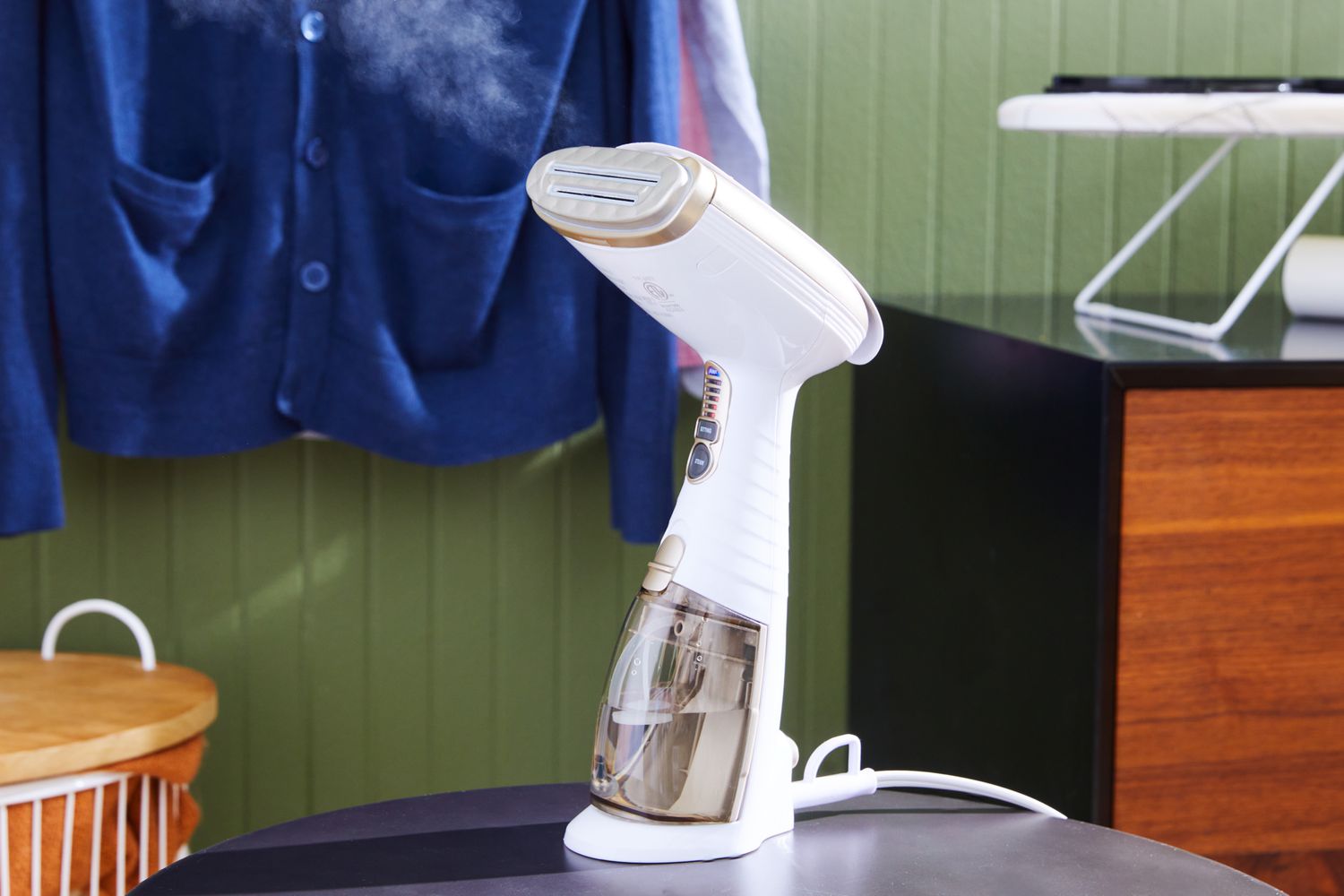 The Conair Turbo ExtremeSteam Hand-Held Fabric Steamer sitting on a table with clothes handing in the back.