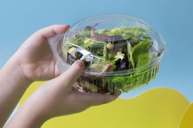 Close up on hands holding a plastic container packed with salad, tomato and baby spinach on vibrant studio background.