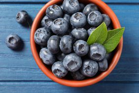 Tasty fresh blueberries with green leaves in bowl on blue wooden table, flat lay