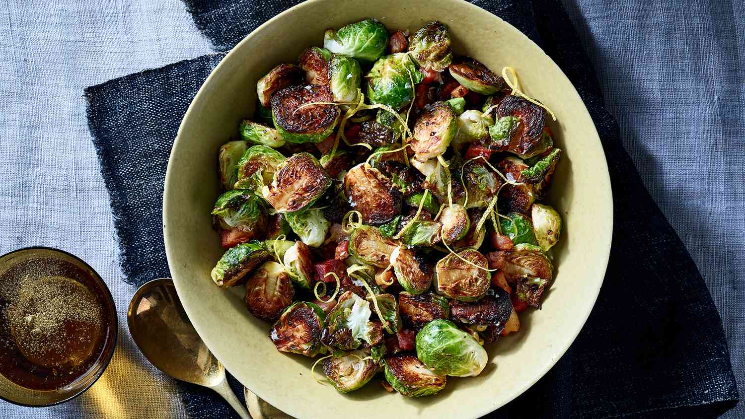 Top View of Roasted Brussels Sprouts With Pancetta Served in a White Bowl on Top of Blue Fabric, with Metal Spoon and Small Metal Bowl of Peppered Oil to the Left