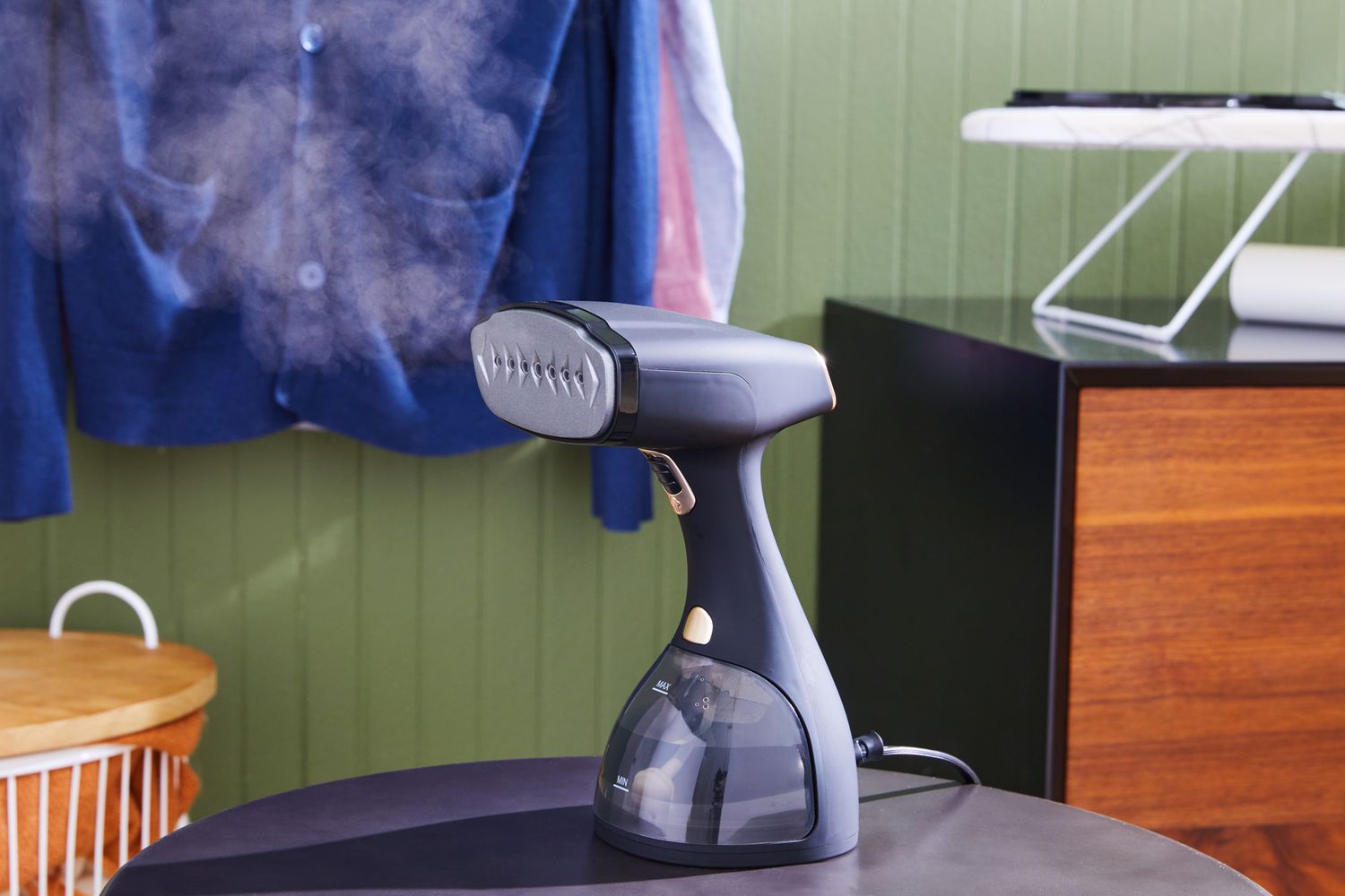The Electrolux LX-15002 Garment Steamer on a counter with steam coming out and clothes hanging in the background. 