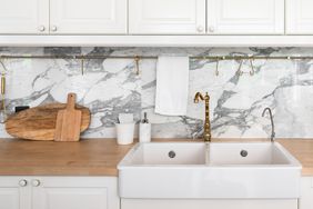 Modern white kitchen interior with white farmhouse sink, gold mirror faucet on wood top with marble backsplash.