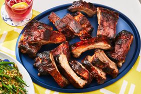 Platter of BBQ ribs set on a yellow print tablecloth with green beans and a red beverage.