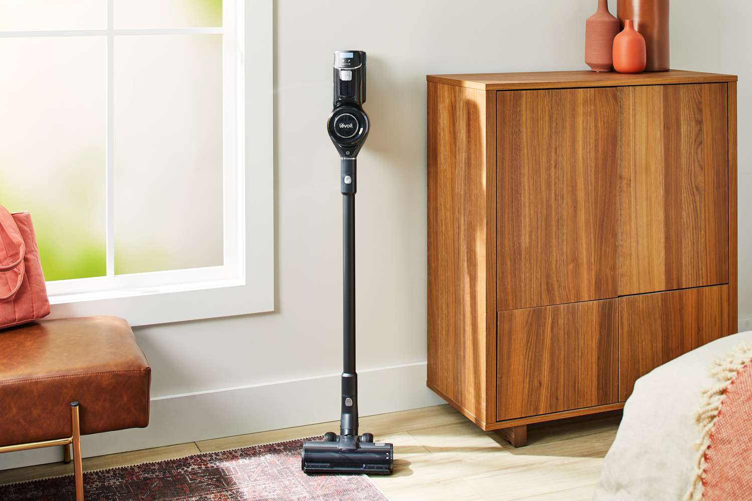 The Levoit VortexIQ 40 Cordless Stick Vacuum leaning against a wall in a bedroom.