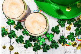 Top view of decorative accessories and drinks for St. Patrick's Day, including shamrock necklaces, gold coins, a green hat, and rainbows