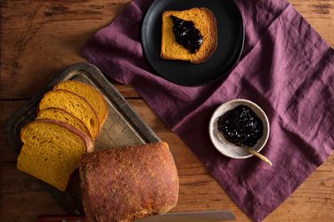 A loaf of pumpkin sandwich bread sliced into five slices, with the rest of the loaf intact. One slice is spread with dark jam and the jam bowl is on the side.