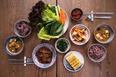 Overhead view of a wooden table full of Korean banchan, purple sticky rice, bowls of soup, and an assortment of lettuce leaves, perilla leaves, carrot sticks, and lengths of quartered cucumber.