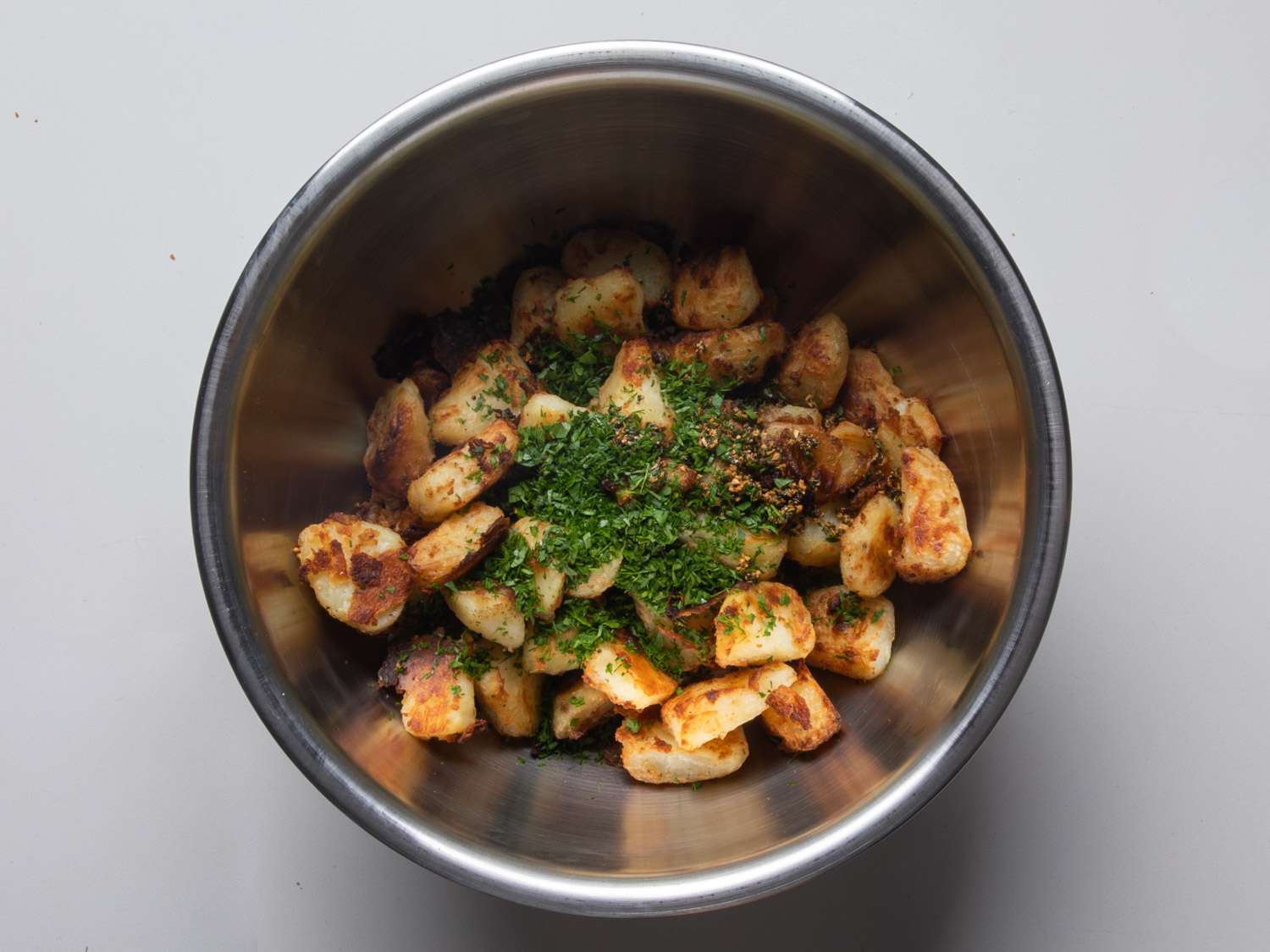 The roasted potatoes in a metal bowl with chopped herbs on top, before the herbs have been mixed in.