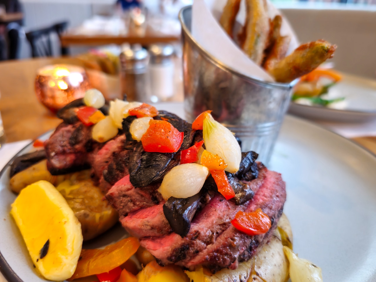 A steak, sliced and topped with pearl onions and peppers, on top of a bed of fingerling potatoes.