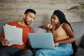 A young couple sitting together on a couch in front of a laptop that's resting on one person's lap looking intently at the screen while the other person holds a document in their hand 