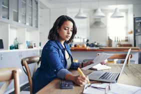 A woman sitting at the table in front of a laptop holding a document and writing with a pencil