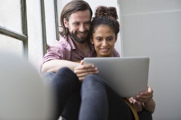 A couple sitting in a windowsill looks at a laptop