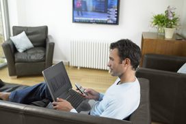 In a living room with the television display on, a man sits with a laptop in his lap while holding a tv remote