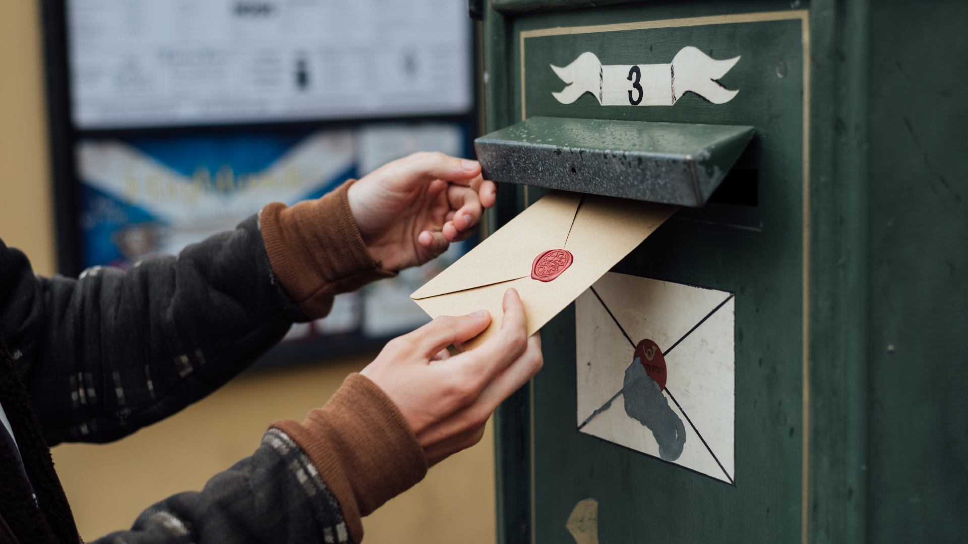 A young woman in a black and brown sweater is placing a letter into a large, slightly weathered, green mailbox on a tree-lined sidewalk.