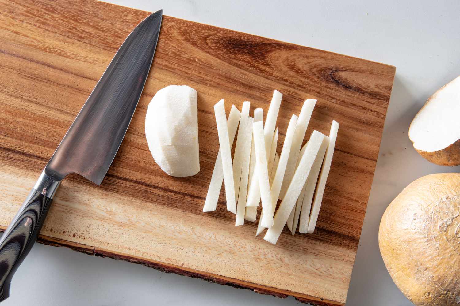A cutting board with a knife and a peeled piece of jicama, cut into thin matchsticks 
