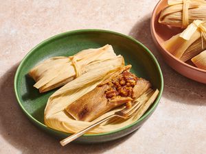 A pecan pie tamale, cut open showing a pecan pie filling, in a green bowl, with a larger bowl of pecan pie tamales in the background