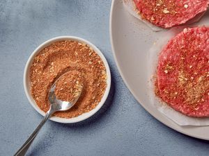 A small bowl of easy burger seasoning next to a plate with two raw burger patties, topped with seasoning