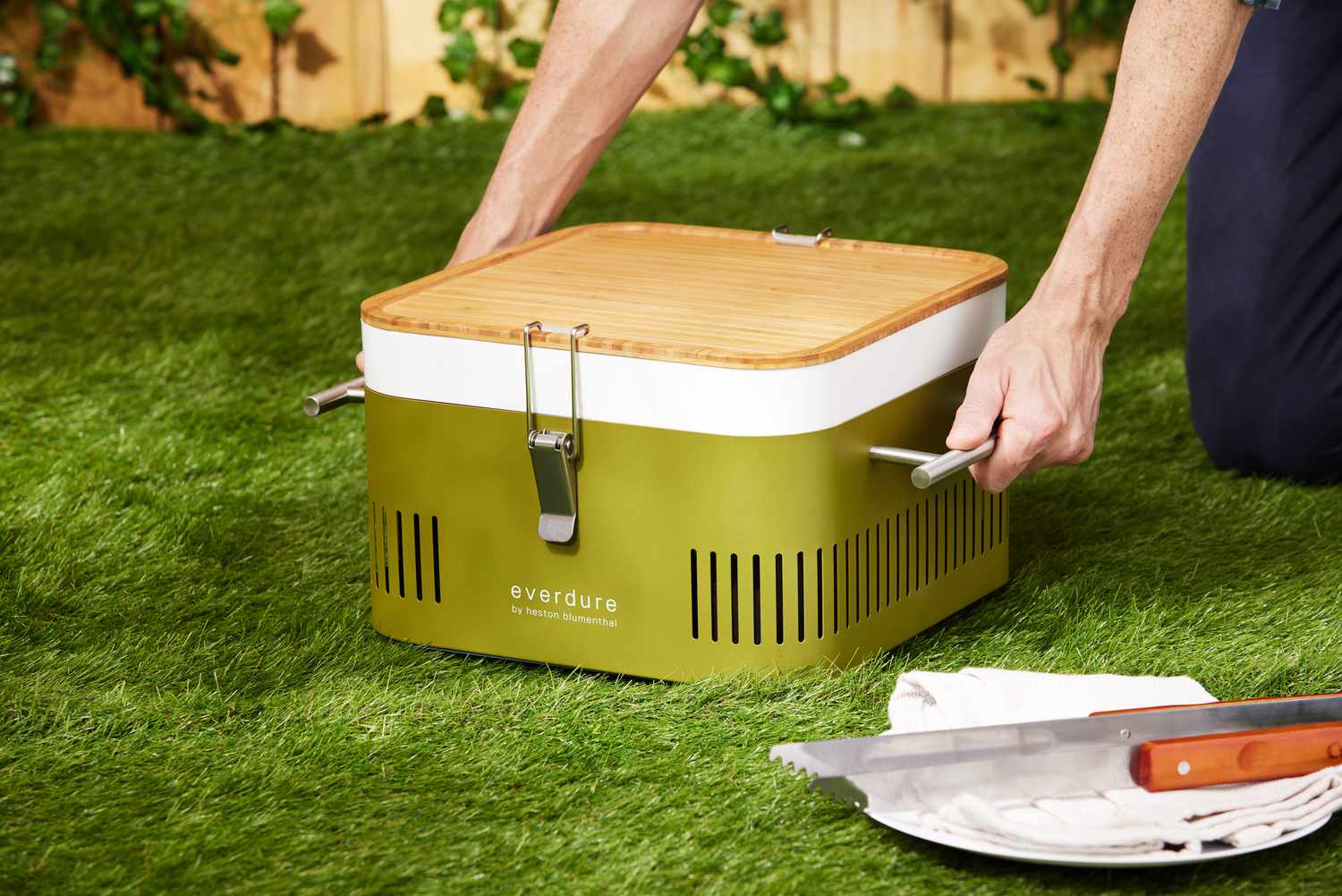 Person lifting the Everdure Cube Portable Charcoal Grill from grass