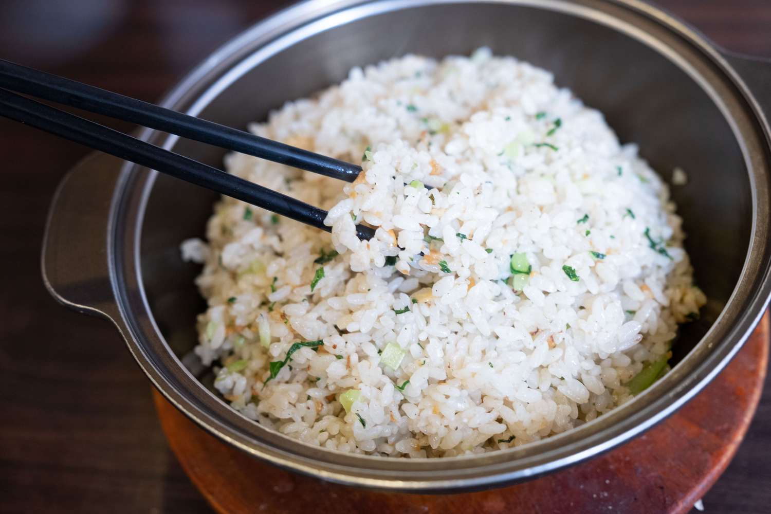 A bowl of vegetable white rice with veggies in the rice. There are two black chopsticks sticking out of the bowl and it sits on a dark wooden table.