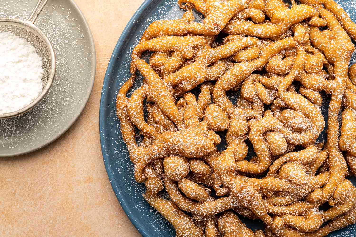 Pancake mix funnel cake lightly dusted with confectioners' sugar on dark-blue serving plate with more powdered sugar in a sieve nearby