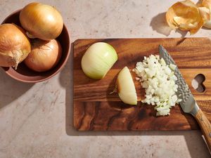 A cutting board with a peeled onion being diced with a knife