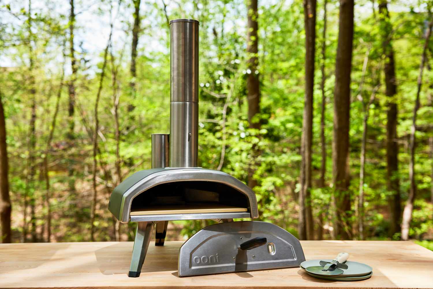 Ooni Fyra 12 Portable Wood-Fired Outdoor Pizza Oven displayed on table in front of woods