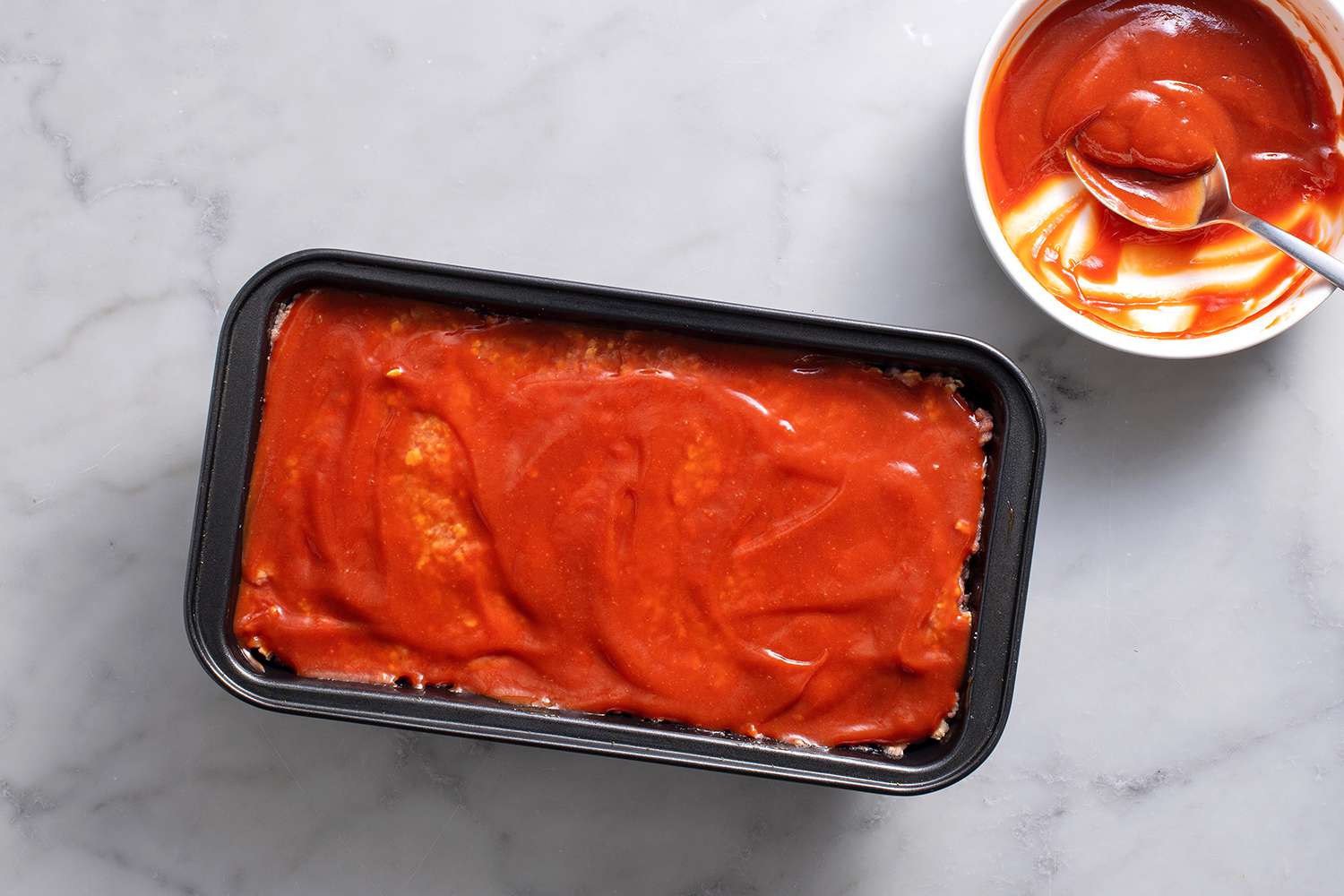 Half of the ketchup glaze evenly spread on raw meatloaf in loaf pan